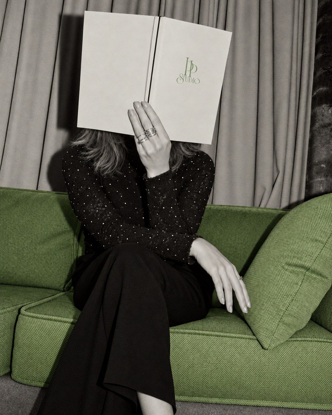 Pia Pucciarello on a green sofa in black and white, her face hidden behind an open book bearing the sage PP Studio logo